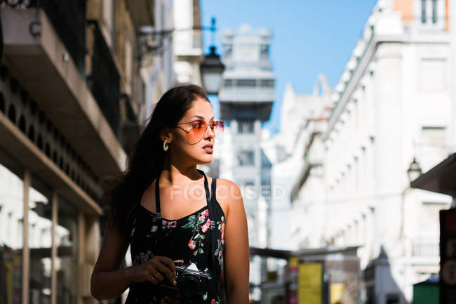 Selbstbewusste schöne Frau im Sommerkleid hält eine Fotokamera in der Hand, während sie auf der malerischen sonnigen Stadtstraße in Lissabon steht, Portugal — Stockfoto
