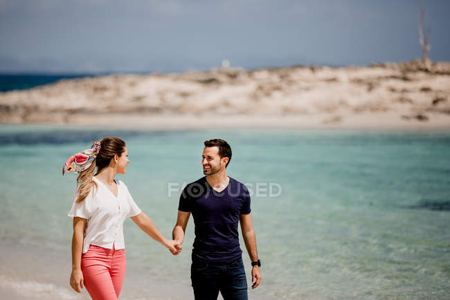 Усміхнений чоловік і жінка йдуть уздовж берега моря, тримаючись за руки і дивлячись один на одного з любов'ю на розмитому фоні — стокове фото
