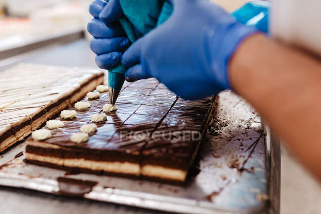 Primer plano empleado anónimo en guantes apretando crema en la parte superior de pasteles de chocolate fresco en bandeja en la panadería - foto de stock