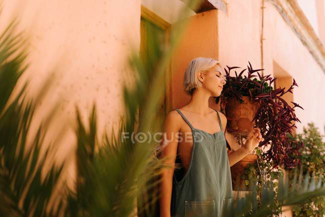 Bella giovane femmina con i capelli biondi corti chiudendo gli occhi e appoggiata al muro mentre in piedi sulla vecchia porta di legno sulla strada della città nella giornata di sole — Foto stock