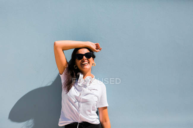 Bella donna in abito casual e cuffie sul collo in piedi con le mani in alto accanto al muro blu di costruzione sulla strada della città — Foto stock