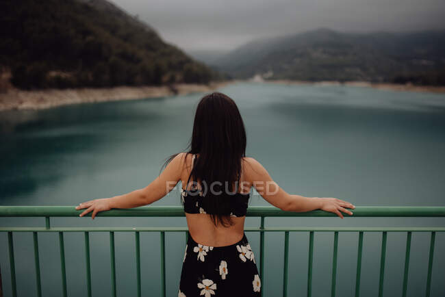 De derrière mince brune en robe dos ouvert appuyé sur la main courante du pont à travers le lac calme à vallée pittoresque de montagne — Photo de stock