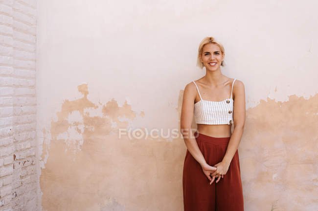 Giovane donna in top alla moda e pantaloni sorridente e guardando la fotocamera mentre in piedi contro muro edificio squallido — Foto stock