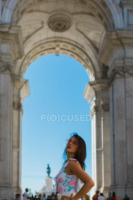 Vista lateral de la joven mujer feliz en gafas de sol de pie junto al majestuoso arco en la calle de la ciudad en Lisboa, Portugal - foto de stock