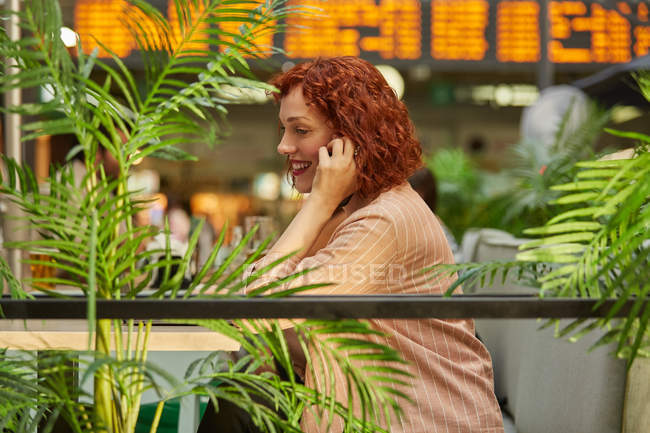 Веселая молодая женщина, работающая на ноутбуке в кафе и разговаривающая по телефону — стоковое фото