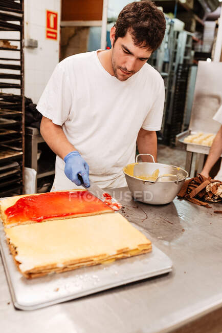Crop man in white uniform holding tray with freshly glazed sweet cakes while working on blurred background of bakery kitchen — Stock Photo