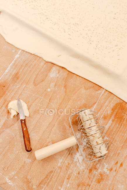 De arriba pequeño cuchillo y rodillo de espiga colocado cerca de la masa fresca delgada en la mesa de madera en la cocina - foto de stock