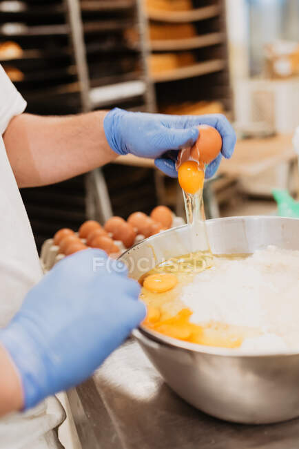 Confiseur anonyme dans des gants bleus brisant l'oeuf de poulet frais dans un bol avec des ingrédients tout en préparant la pâte à pâtisserie dans la cuisine de boulangerie — Photo de stock