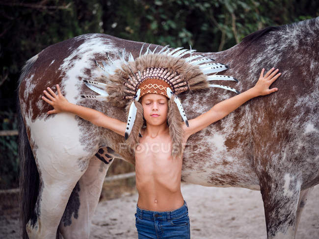 Criança cuidadosa em chapéu de penas indiano com olhos fechados apoiados em garanhão com as mãos estendidas no rancho — Fotografia de Stock