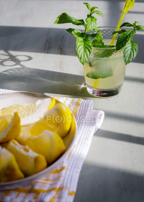 Coloridos trozos de limón en el plato compuesto con refrescante bebida de limón con hojas de menta en la mesa de la cocina de madera - foto de stock