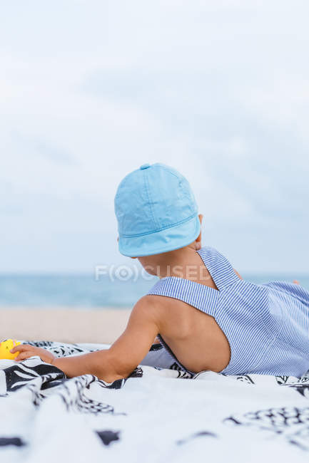 Vista trasera de un bebé con una gorra en la playa junto a sus patos de goma - foto de stock