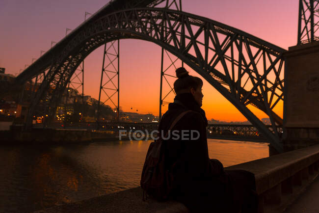 Vista trasera de la turista mirando hacia otro lado mientras está sentada en la frontera del terraplén de la ciudad cerca del puente durante la puesta del sol en Oporto, Portugal - foto de stock