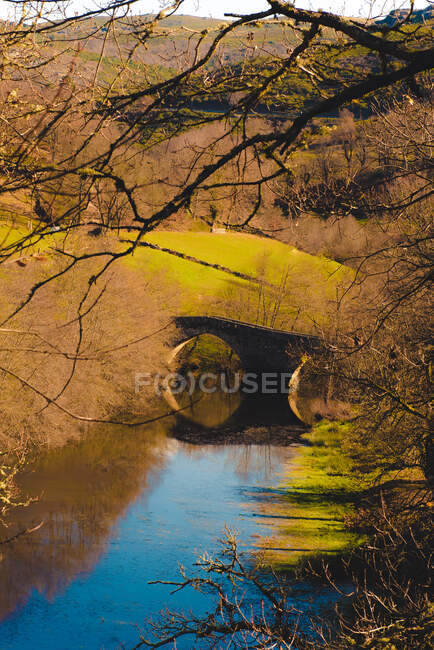 Выдержанный каменный мост через спокойную реку в солнечный день в живописной осенней сельской местности — стоковое фото