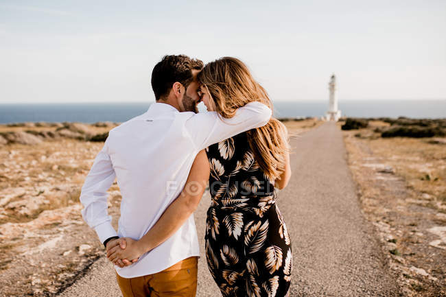 Vista trasera de la tierna pareja abrazándose en el camino de asfalto al faro y la orilla del mar en verano - foto de stock