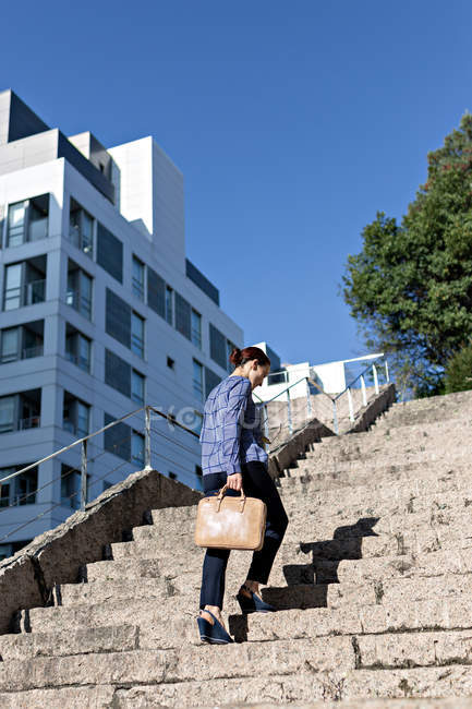 Low angle of elegant entrepreneur with stylish briefcase walking up rough steps on sunny day on city street — Stock Photo