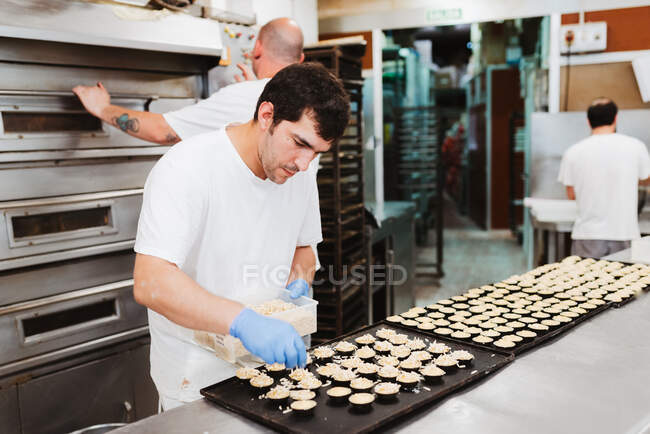 Homem adulto de uniforme branco e luvas azuis colocando flocos de coco frescos em deliciosa confeitaria enquanto estava perto da mesa e trabalhando na padaria — Fotografia de Stock