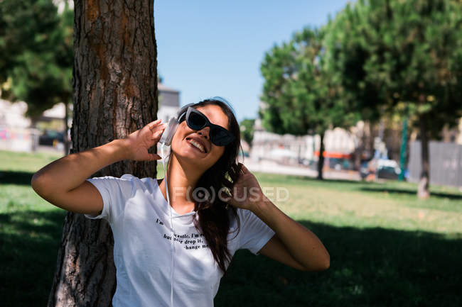 Friedliche Frau mit Sonnenbrille und Kopfhörer, die Musik hört, während sie im Park auf Gras sitzt — Stockfoto