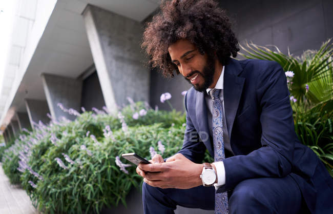 Joyful african american man in suit sitting outside and messaging — Stock Photo