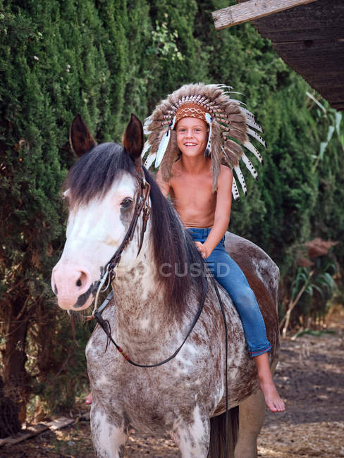 Счастливый мальчик в настоящей индийской шляпе с перьями верхом на лошади в парке — стоковое фото