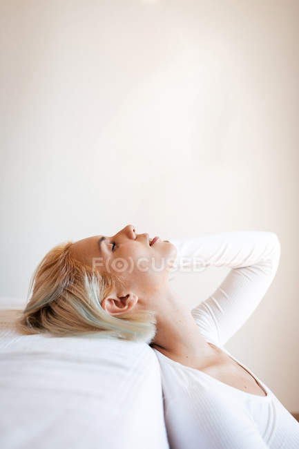 Seitenansicht der schönen blonden Frau, die die Augen schließt und sich zu Hause auf eine bequeme Matratze an die weiße Wand lehnt — Stockfoto