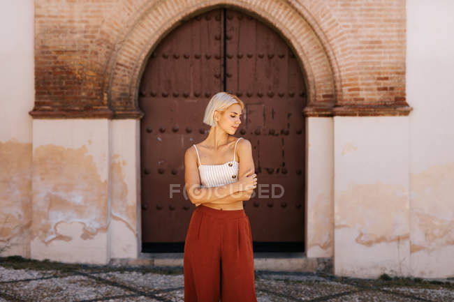 Jovem mulher atraente em roupas elegantes braços dobráveis e fechar os olhos enquanto está de pé contra o edifício antigo com portão gasto — Fotografia de Stock