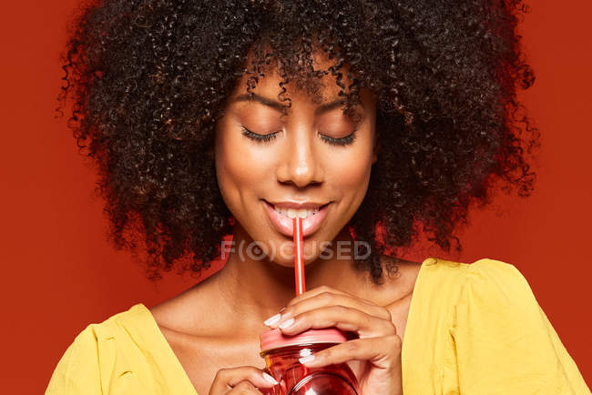Vista de close-up da mulher afro-americana segurando frasco vermelho com palha e apreciando bebidas no fundo vermelho — Fotografia de Stock