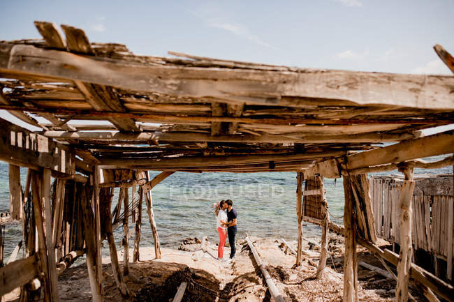 Вид на романтическую пару, обнимающуюся на побережье возле деревянного навеса летом — стоковое фото