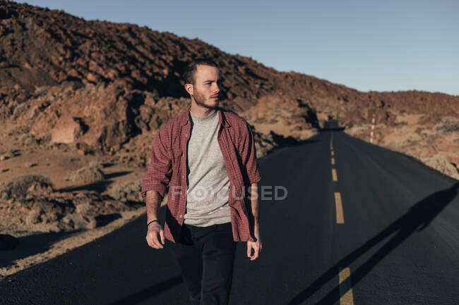 Молодой красивый мужчина в повседневной одежде, прогуливаясь по пустынной горной дороге на Тенерифе во время заката, смотрит в сторону — стоковое фото