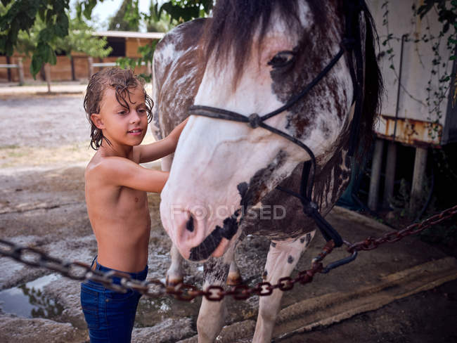 Happy shirtless kid with curly wet hair embracing horse side on ranch — Stock Photo