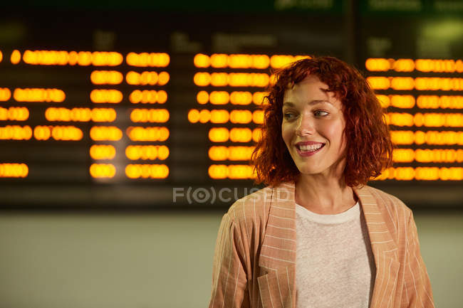 Alegre vermelho encabeçado jovem mulher de pé perto de horário na estação ferroviária e olhando para longe — Fotografia de Stock