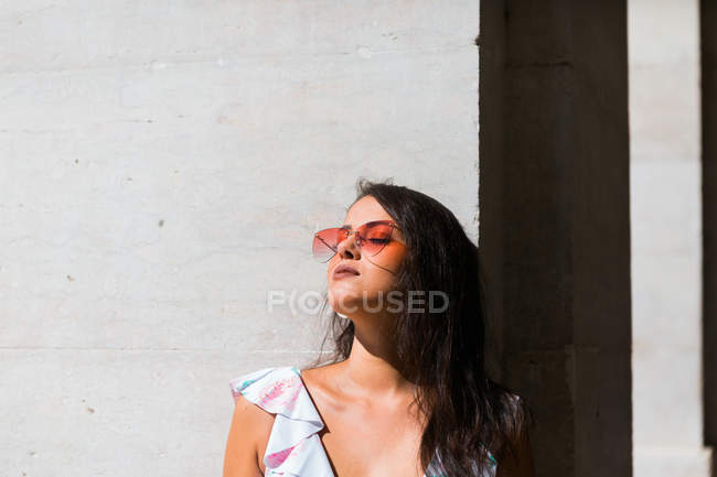 Peaceful gorgeous woman in trendy outfit and shiny sunglasses standing on white wall on scenic street — Stock Photo