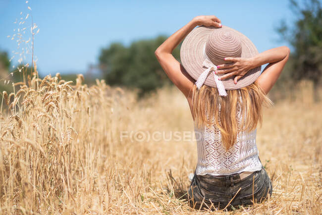 Back view of slim female touching hat while sitting on dry grain grass in meadow on sunny summer day in nature — Stock Photo