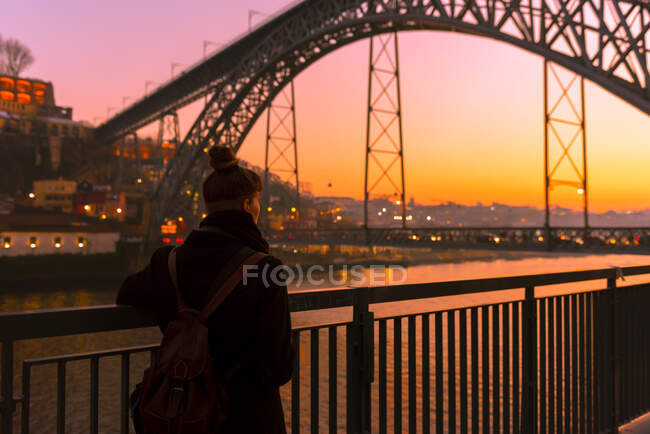 Rückansicht einer Touristin, die in der Nähe des Stadtdamms in der Nähe einer Brücke steht und bei Sonnenuntergang in Porto, Portugal, wegschaut — Stockfoto