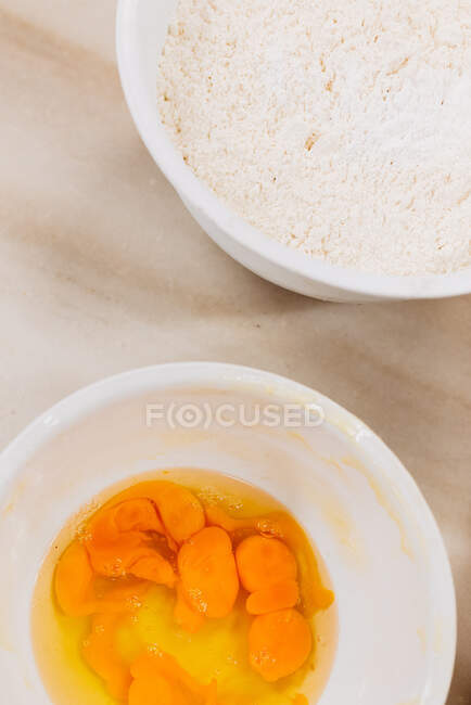 Сверху миски с сырыми яйцами и пшеничной мукой помещаются на кухонный стол во время приготовления выпечки — стоковое фото