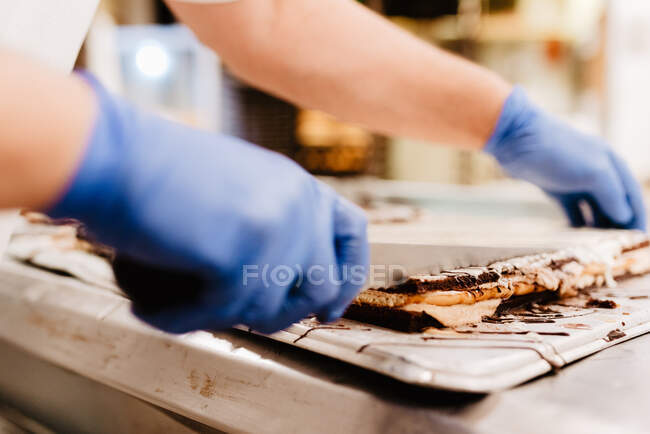 Anonymous cook in gloves cutting delicious layered cake on table in bakery kitchen — Stock Photo