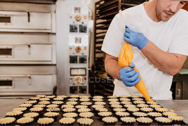 Pastelero anónimo en guantes de látex exprimiendo sabrosa jalea dulce de la bolsa en pequeñas cajas de pastelería en el fondo borroso de la cocina de panadería - foto de stock