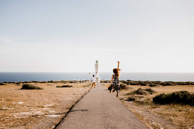 Обратный вид взволнованных женщин и мужчин, прыгающих по дороге к маяку и морю в летний солнечный день — стоковое фото
