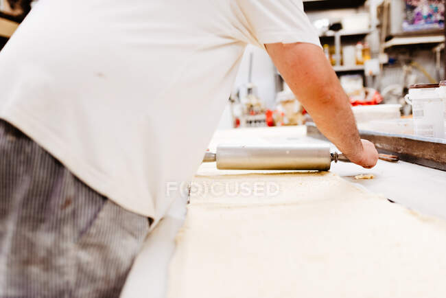 Homem sobrepeso anônimo em uniforme rolando massa macia fresca na mesa enquanto cozinha pastelaria — Fotografia de Stock