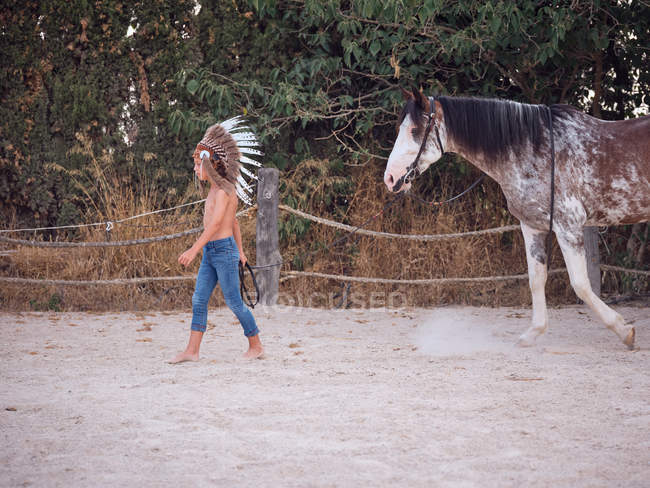 Side view of calm kid wearing feather Indian war bonnet and walking shirtless on sandy farm, leading horse behind — Stock Photo