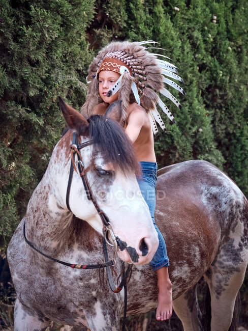 Serio ragazzo in autentico indiano piuma cappello equitazione cavallo in parco — Foto stock