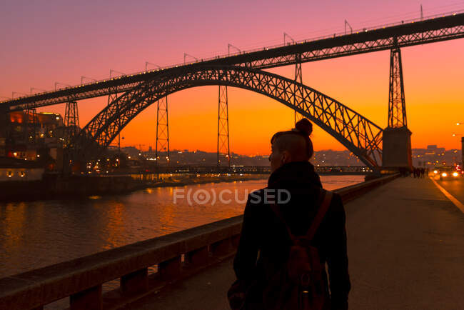 Обратный вид на неузнаваемую женщину-туристку, идущую рядом с городской набережной возле моста во время заката в Порту, Португалия — стоковое фото