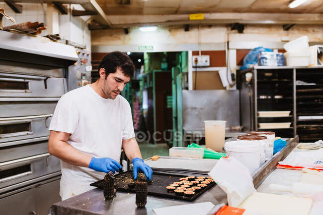 Cook squeezing fresh pastry dough on tray with paper while working on blurred background of bakery — Stock Photo
