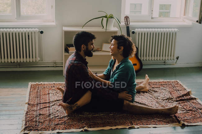 Side view of tender couple in casual clothes cuddling and smiling while sitting on carpet on floor at home — Stock Photo