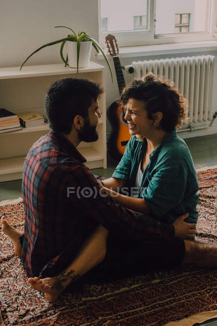 Side view of tender couple in casual clothes cuddling and smiling while sitting on carpet on floor at home — Stock Photo