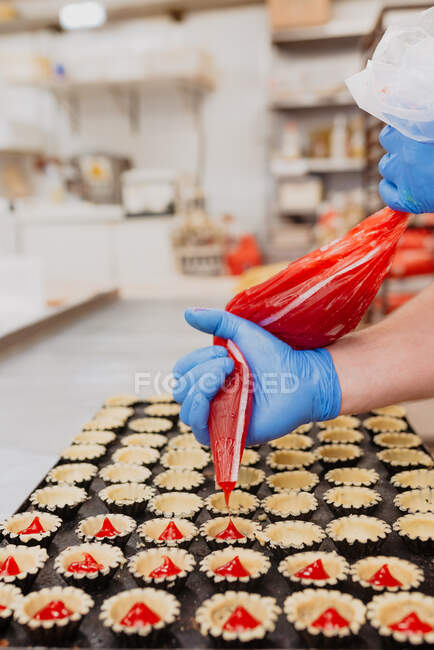 Confiseur anonyme en gants de latex pressant une délicieuse gelée sucrée du sac dans de petites caisses de pâtisserie sur fond flou de cuisine de boulangerie — Photo de stock