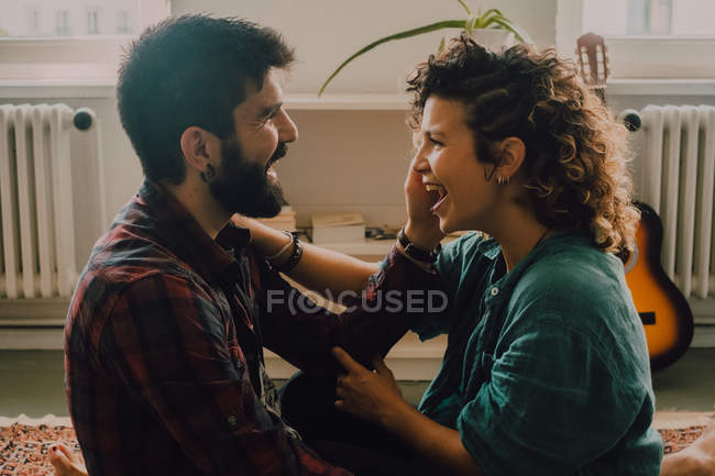 Vista lateral de casal macio abraçando e sorrindo enquanto sentado no chão em casa e tocando uns aos outros rostos — Fotografia de Stock