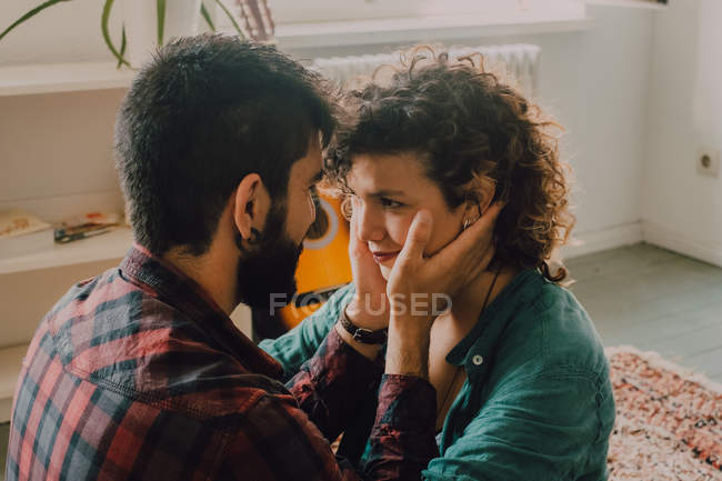 Vista lateral de casal macio abraçando e sorrindo enquanto sentado no chão em casa e tocando uns aos outros rostos — Fotografia de Stock