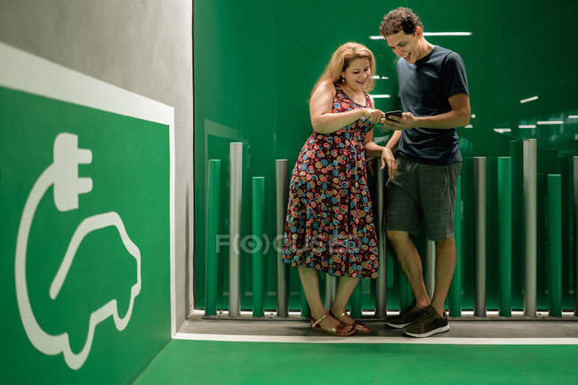 Smiling joyful couple waiting for electric car to charge and sharing mobile phone while standing in light hall — Stock Photo