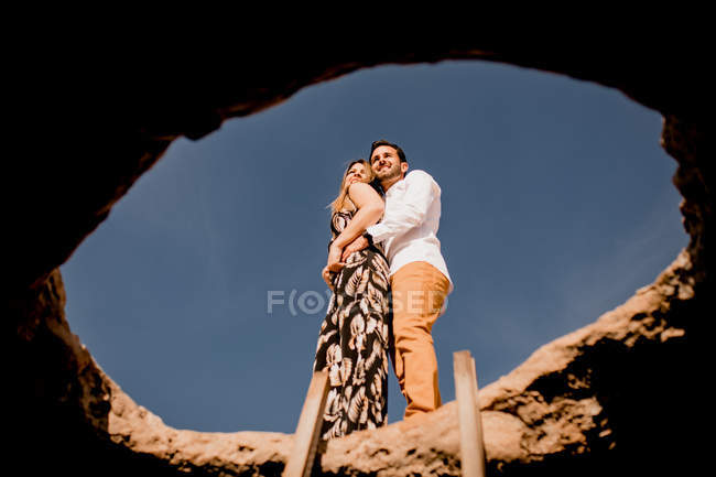 De baixo de amar o jovem e a mulher de vestido abraçando na pedra bem contra o céu azul claro — Fotografia de Stock