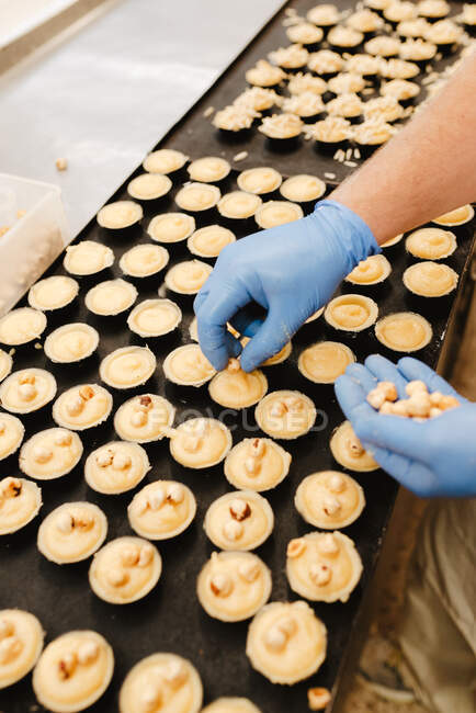 Von oben anonymer Mitarbeiter im Handschuh, der frische Haselnüsse auf leckeres süßes Gebäck auf Tablett legt, während er in der Bäckerei arbeitet — Stockfoto